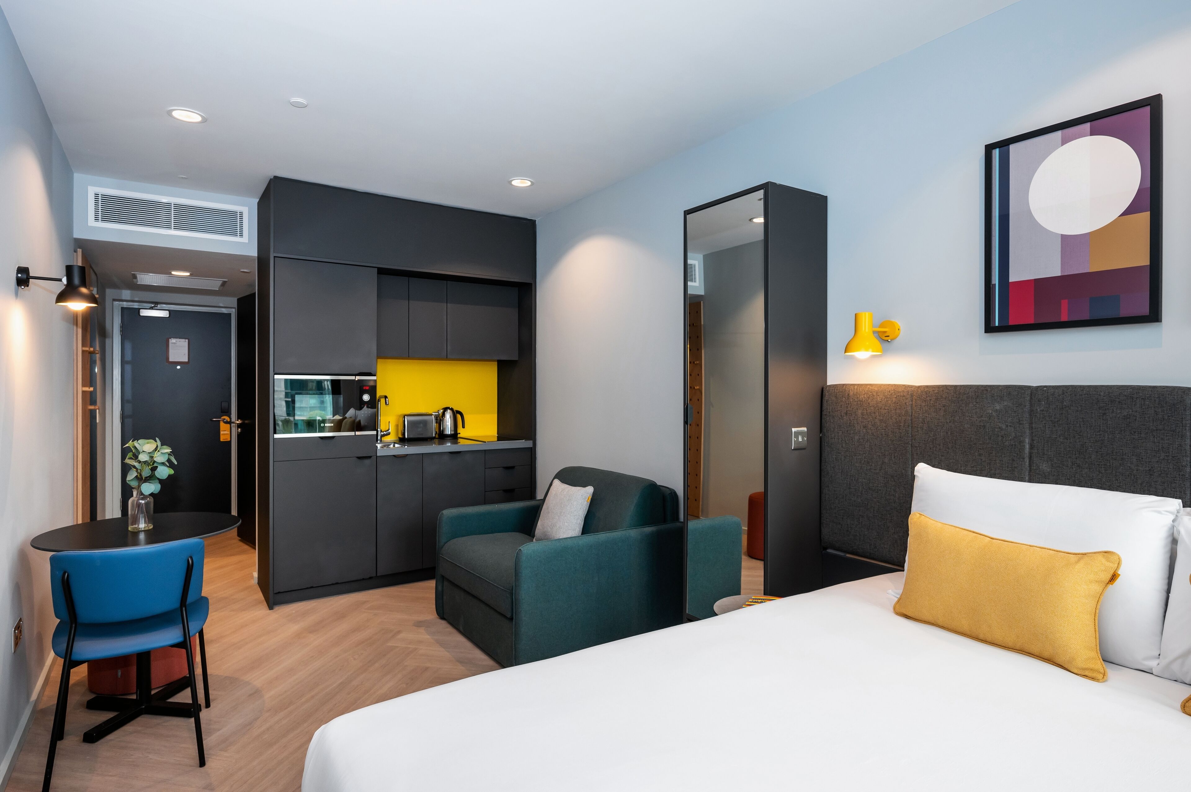 A modern hotel room with a bed, a desk, a chair, and a kitchenette.