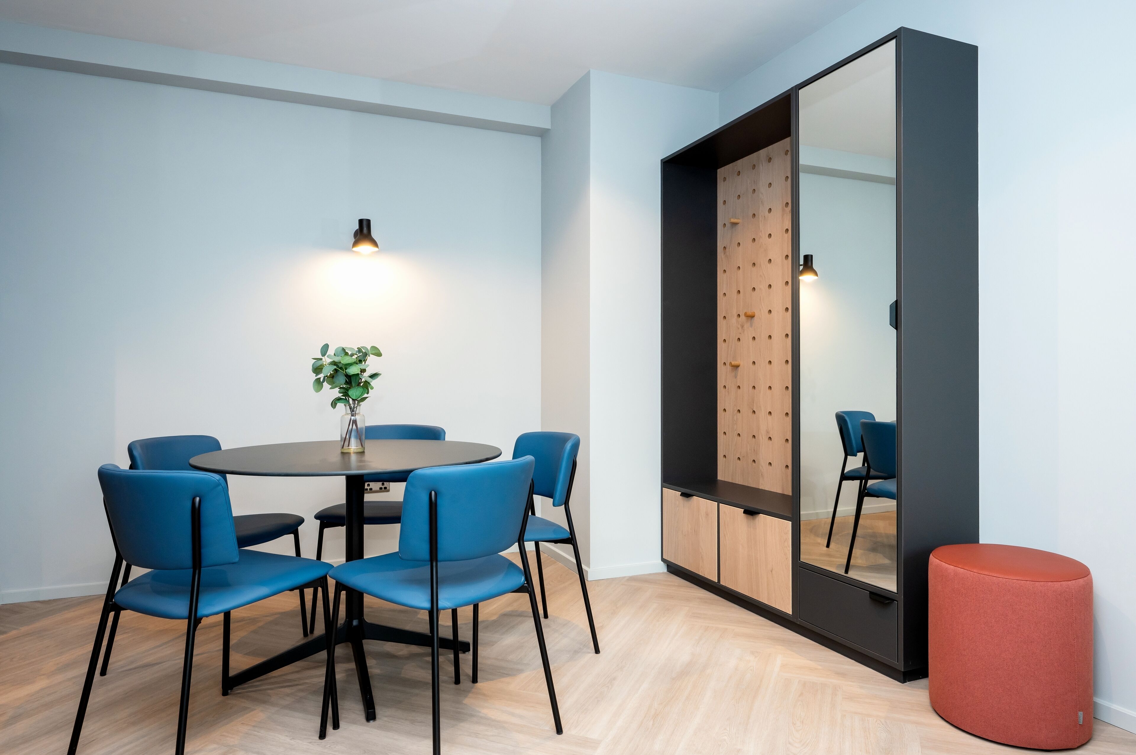 Ein modernes Zimmer mit einem runden Tisch, blauen Stühlen, einem roten Pouf und einem großen Spiegel mit Holzverkleidung.