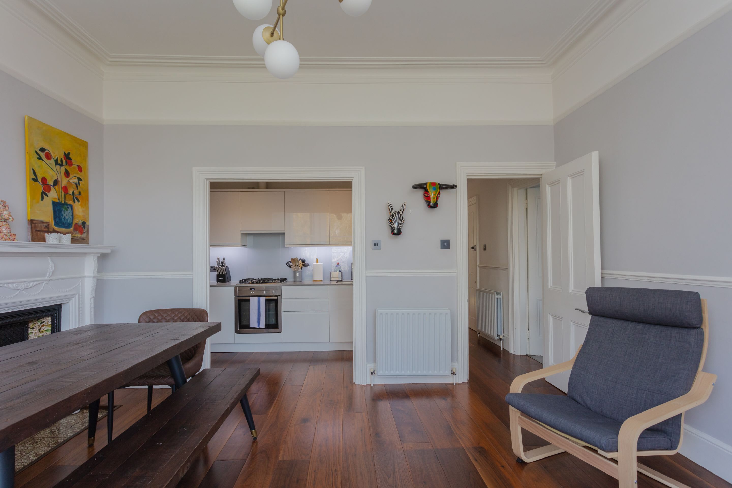 A dining area with a wooden table and benches, a kitchen in the background, and a grey armchair.