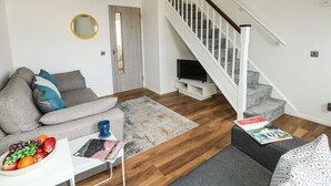 Uma sala de estar moderna com um sofá cinzento, uma mesa de centro com frutas e uma revista, uma escada com grades brancas e uma luminária de teto de madeira.