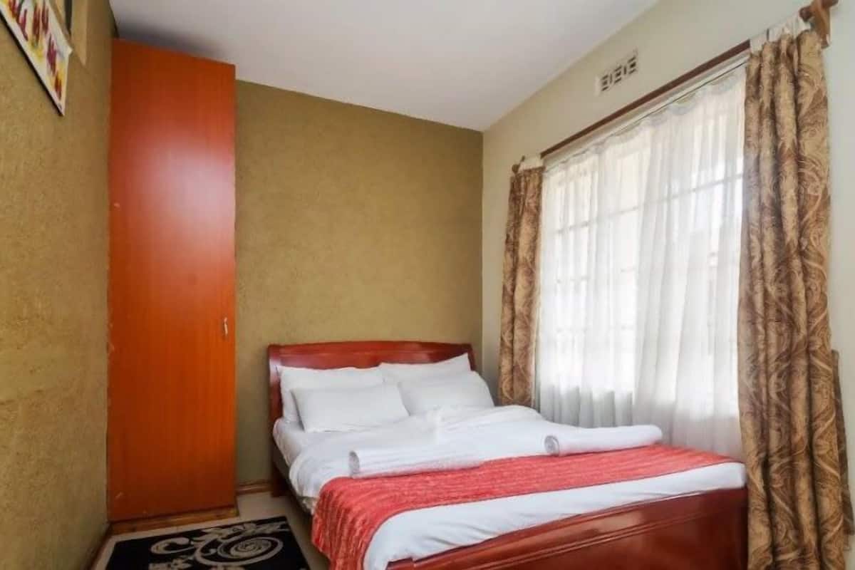 A small, single-bed room with a wooden headboard, a red blanket, and a window with patterned curtains.