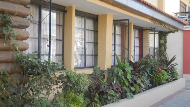 A building with a covered outdoor area, featuring a row of windows with metal bars, a tiled floor, and a variety of plants. - The Stand Leisure Nairobi West (Nairobi)