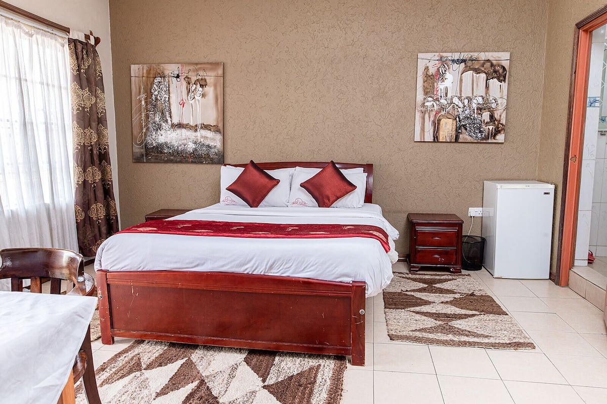 A bedroom with a bed, a nightstand, a chair, a desk, a refrigerator, and two paintings on the wall.