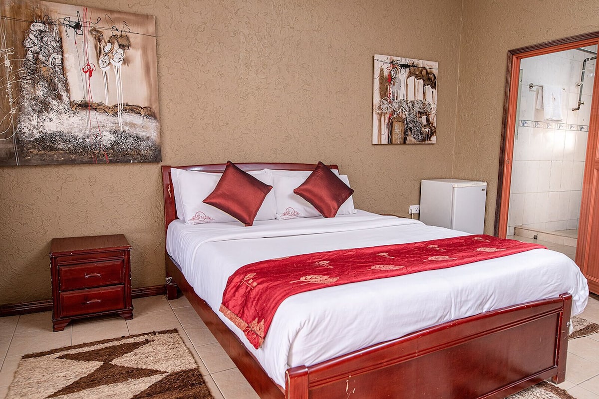 A hotel room with a wooden bed, white bedding, a red throw, a nightstand, a small cabinet, and two paintings on the wall.