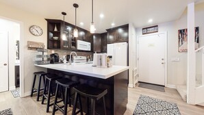 A modern kitchen with dark wood cabinets, a white island with bar stools, a clock, and a welcome sign. - St. Anton 54 Steps to Canyon Lodge, Remodeled, Covered Parking by RedAwning (Mammoth Lakes)