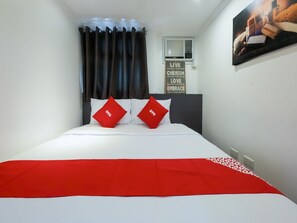 Ein Hotelzimmer mit einem Bett, zwei roten Kissen, einer roten Decke, einem Fenster mit Vorhängen, einer Klimaanlage und einem motivierenden Zitat an der Wand.