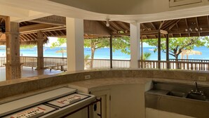 A covered outdoor area with a view of the ocean, wooden pillars, and a tiled counter with a sink.