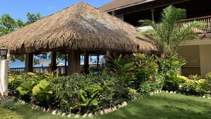 A thatched-roof structure with a view of the sea, surrounded by lush tropical plants and a well-maintained lawn.
