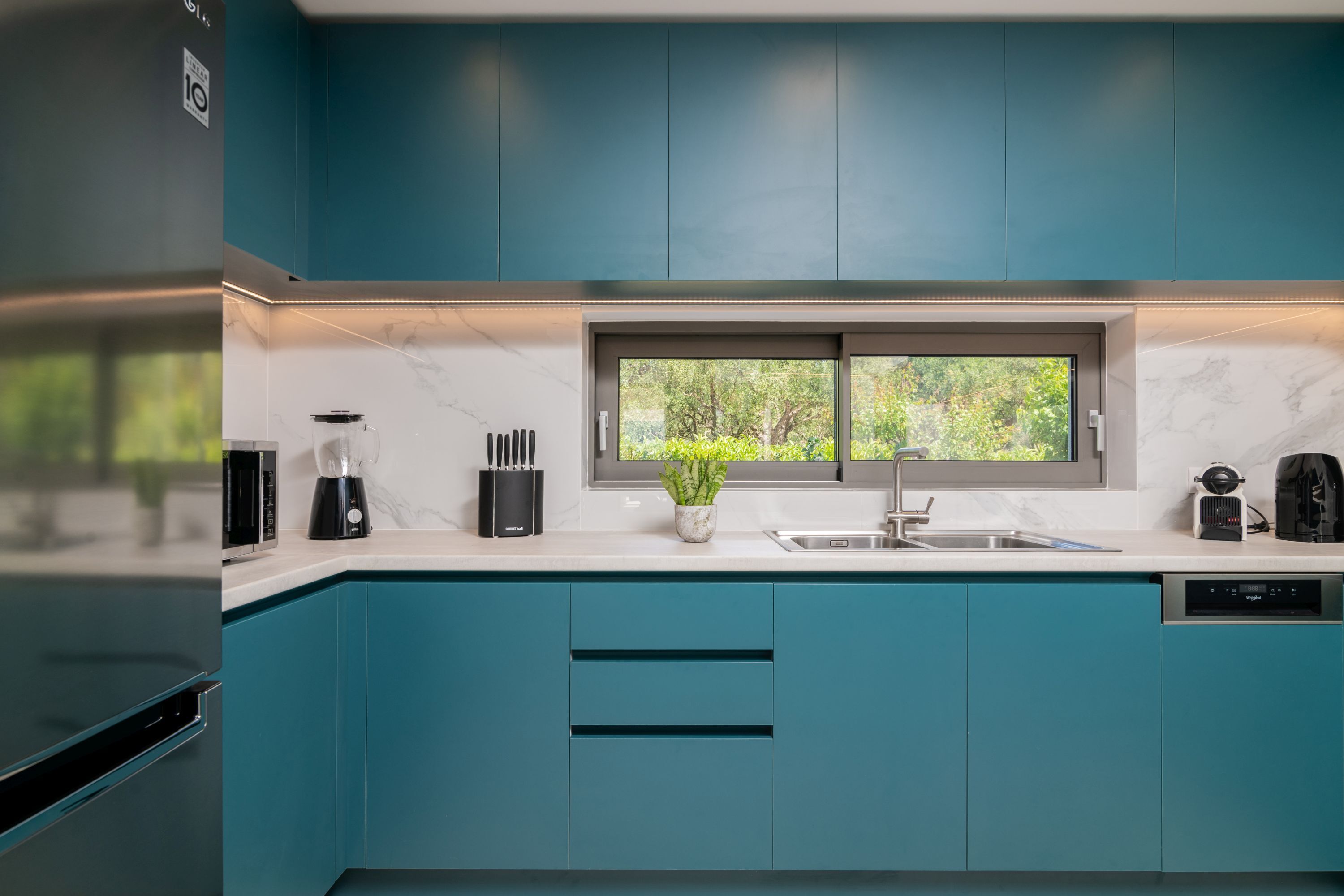 Una cocina moderna con armarios azul verdoso, encimera de mármol, ventana con vistas a la vegetación y lavavajillas.