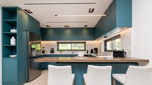 Una cocina moderna con armarios color turquesa, una isla central con taburetes de bar, electrodomésticos de acero inoxidable y un amplio ventanal que proporciona luz natural.
