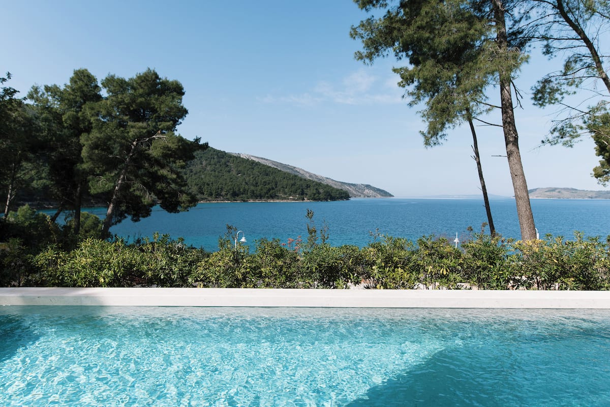 A pool with clear blue water, surrounded by greenery and overlooking a calm sea and a hilly landscape.
