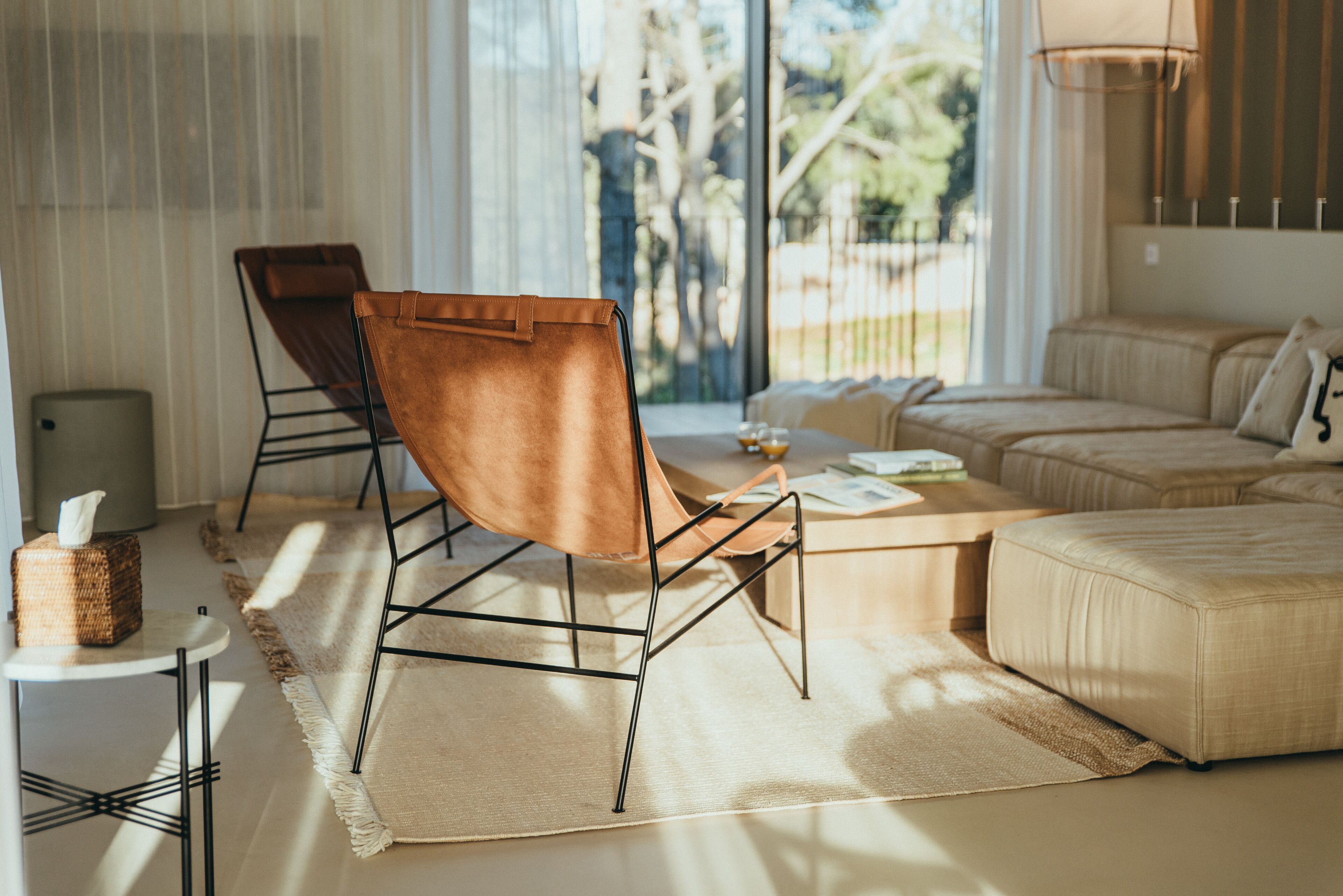 A modern living room with a sofa, two chairs, a coffee table, and a view of trees through large windows.