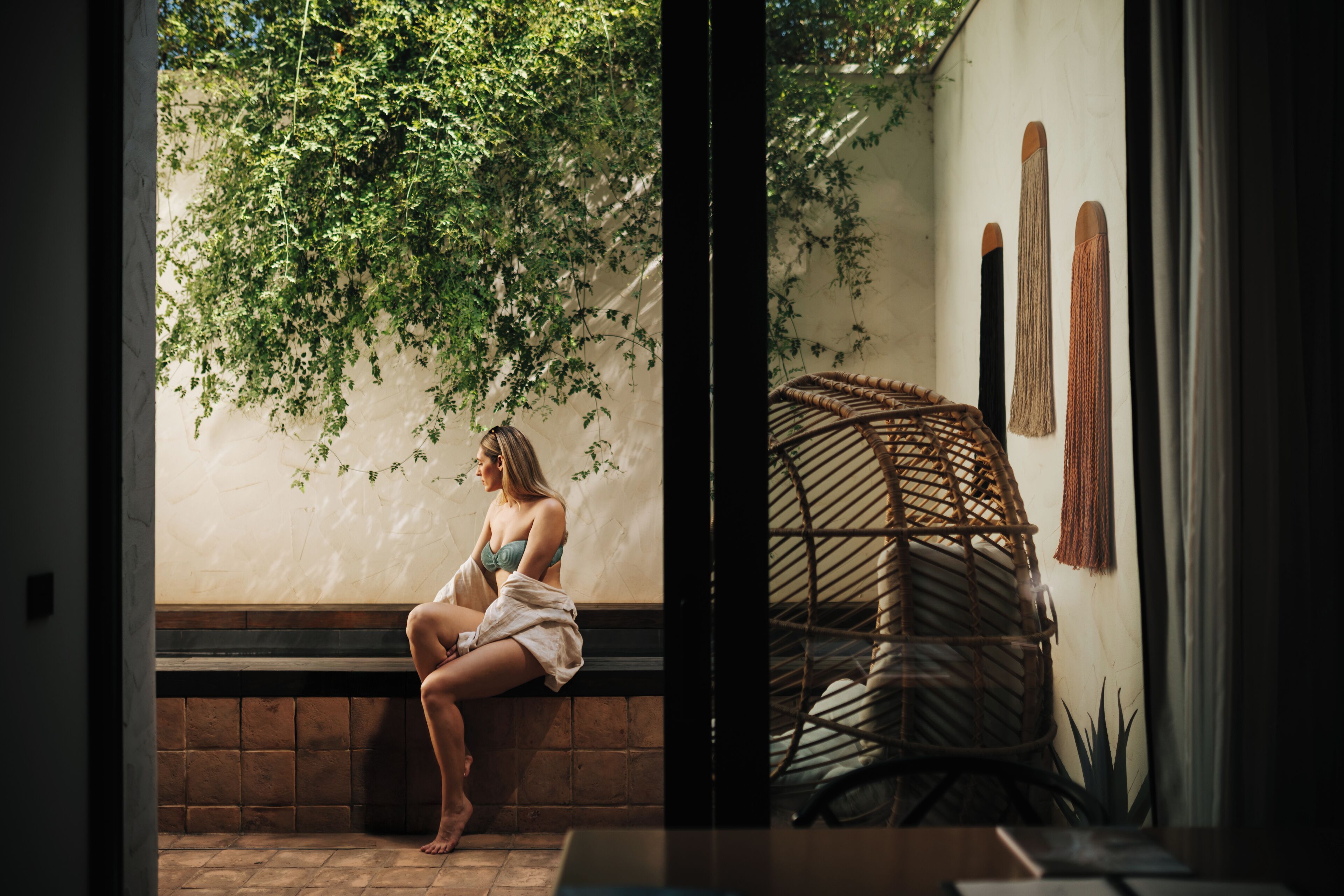 A woman sitting on a bench in a room with a wicker chair and a wall-mounted macrame piece.