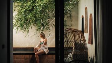 A woman sitting on a bench in a room with a wicker chair and a wall-mounted macrame piece.