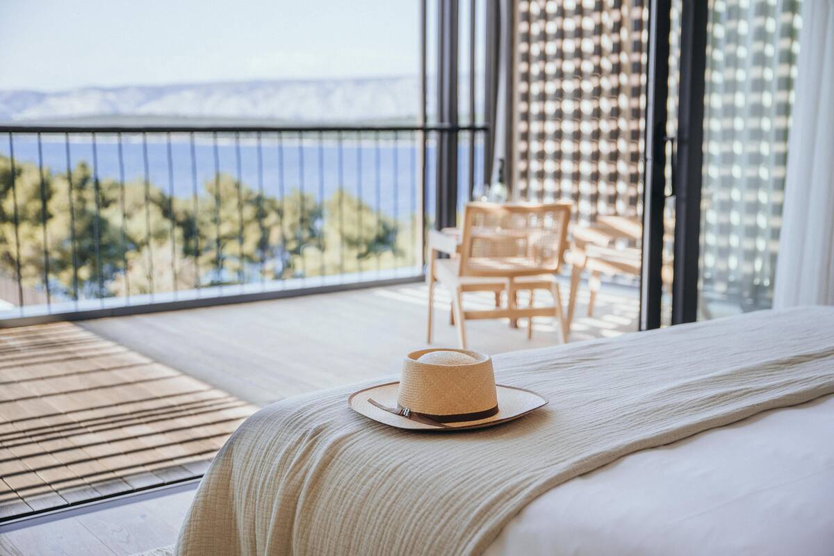 A bedroom with a bed, a straw hat on a bedside table, and a balcony with a view of the sea and trees.