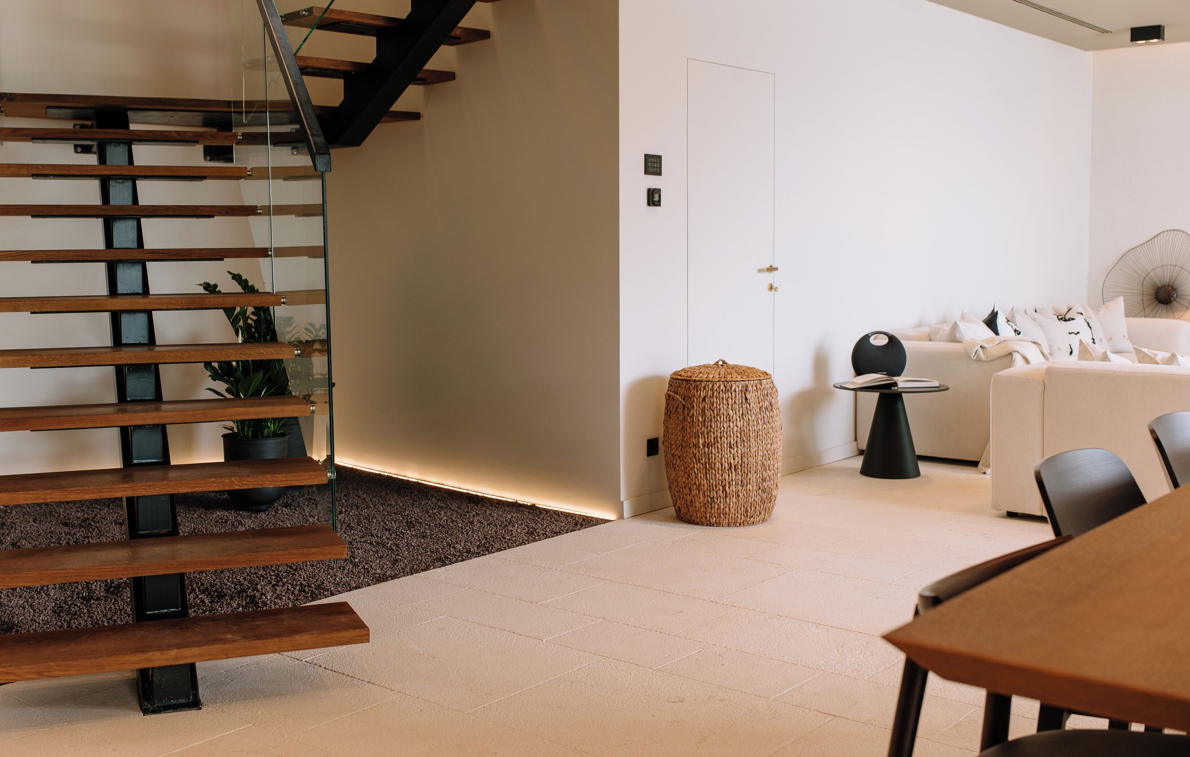 A modern interior with a wooden staircase, a white door, a round woven basket, and a white sofa with black chairs.