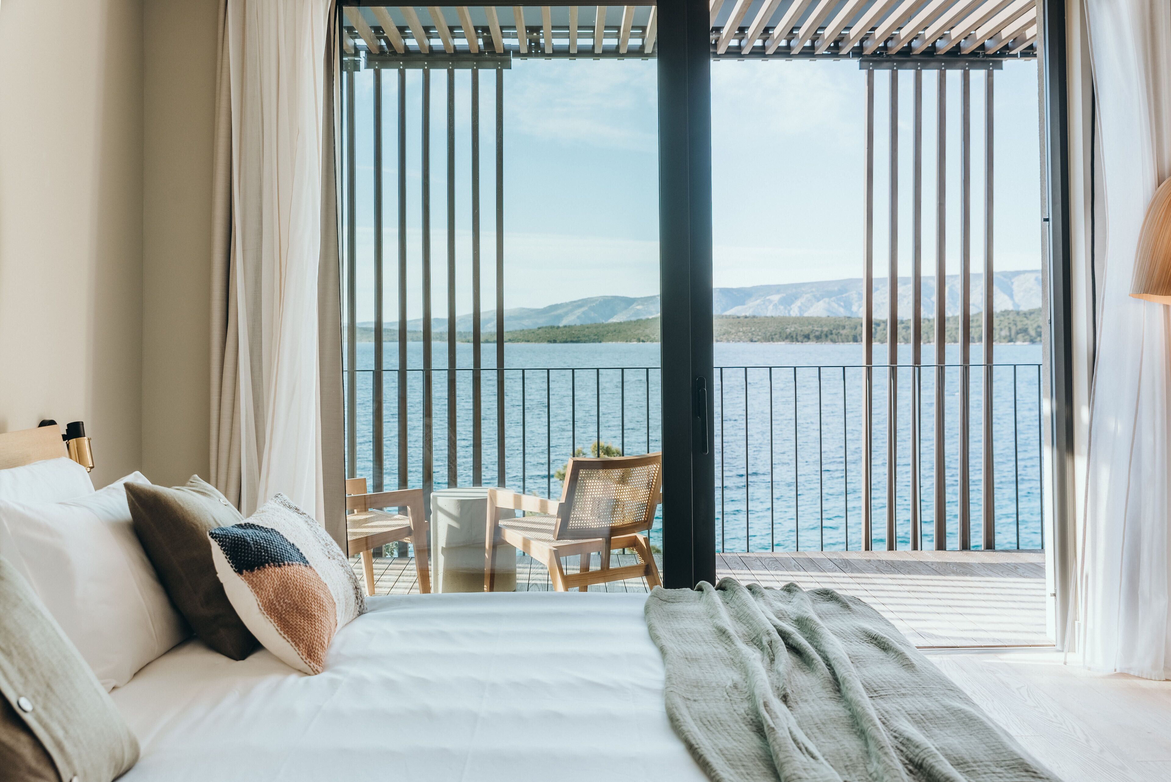 A bedroom with a large window overlooking a body of water and mountains. There is a bed with pillows and a blanket, a chair, and a table.