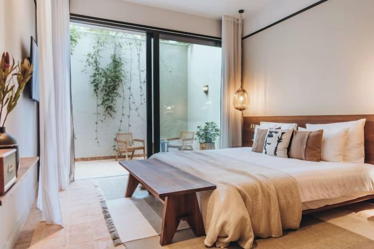 A bedroom with a large bed, a bench, and a view of a garden through a sliding door.