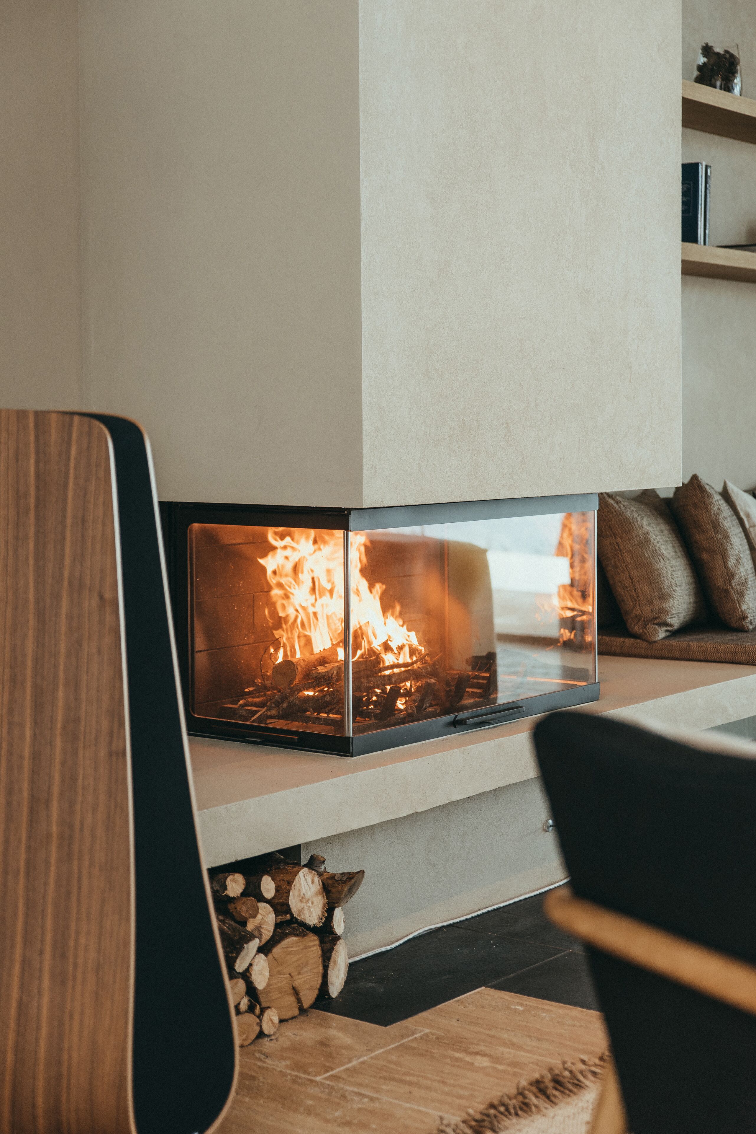 Una chimenea moderna con frente de vidrio, una silla de madera al frente y un montón de leña abajo.