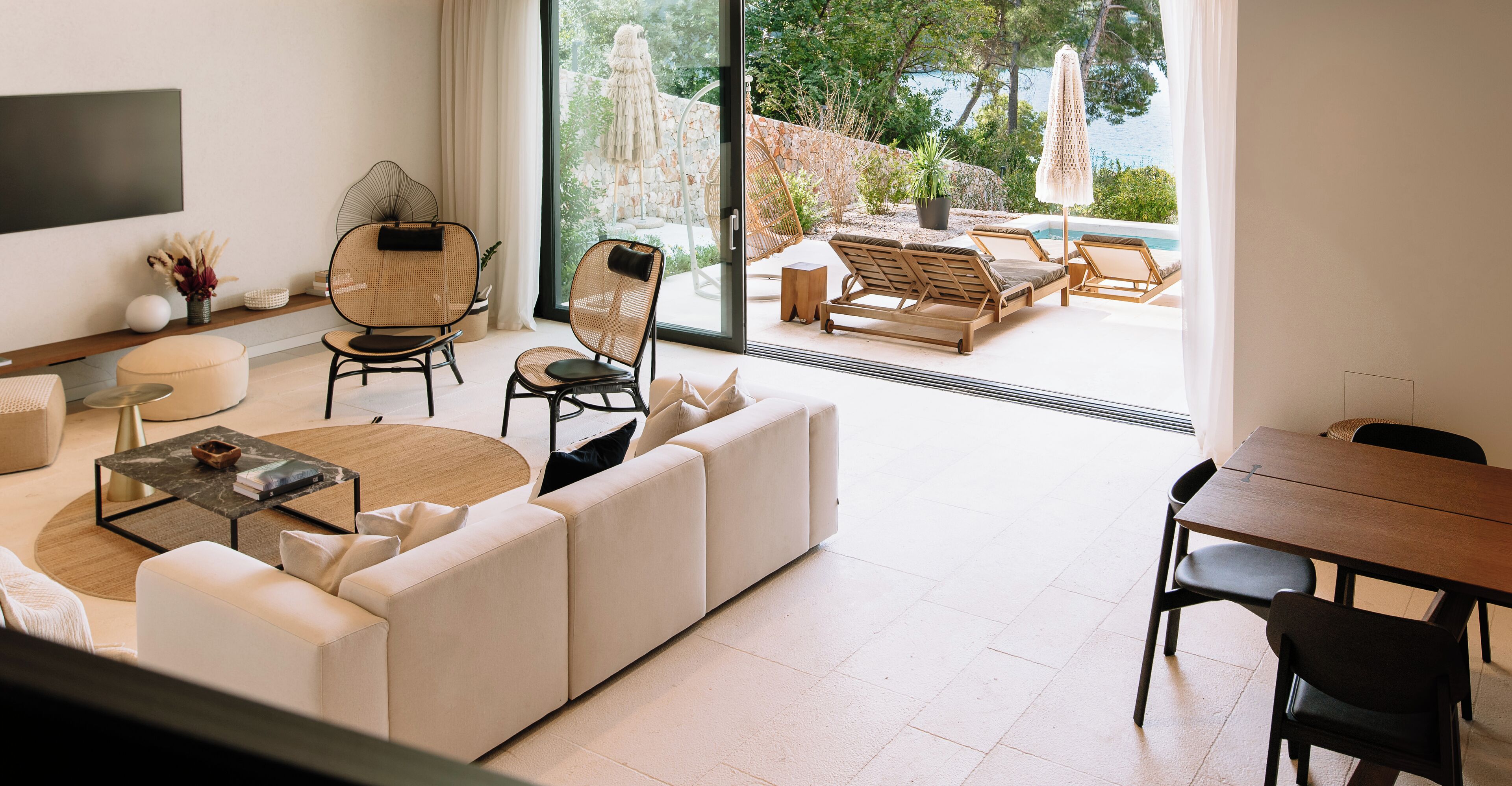 A modern living room with a large sofa, a coffee table, and a dining area with a table and chairs. There is a large window with a view of the outdoors.