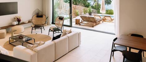 A modern living room with a large sofa, a coffee table, and a dining area with a table and chairs. There is a large window with a view of the outdoors.
