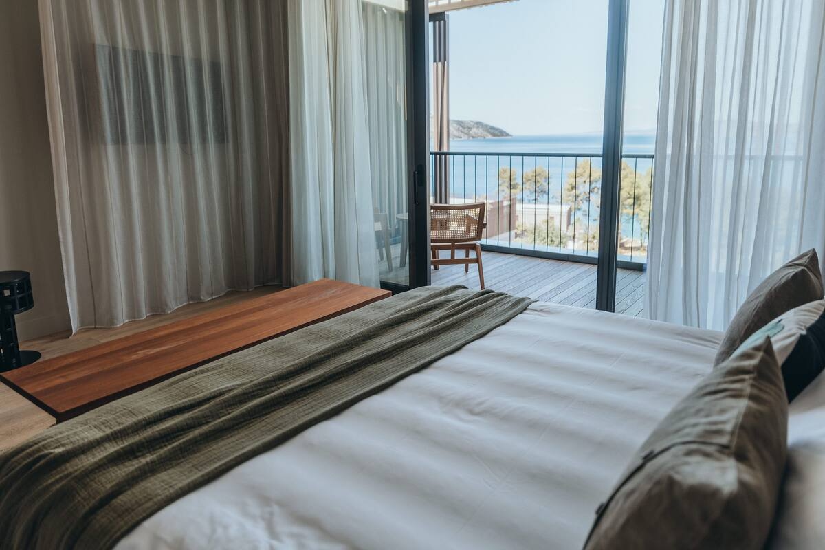 A hotel room with a large bed, a wooden side table, a chair, and a balcony with a view of the sea.