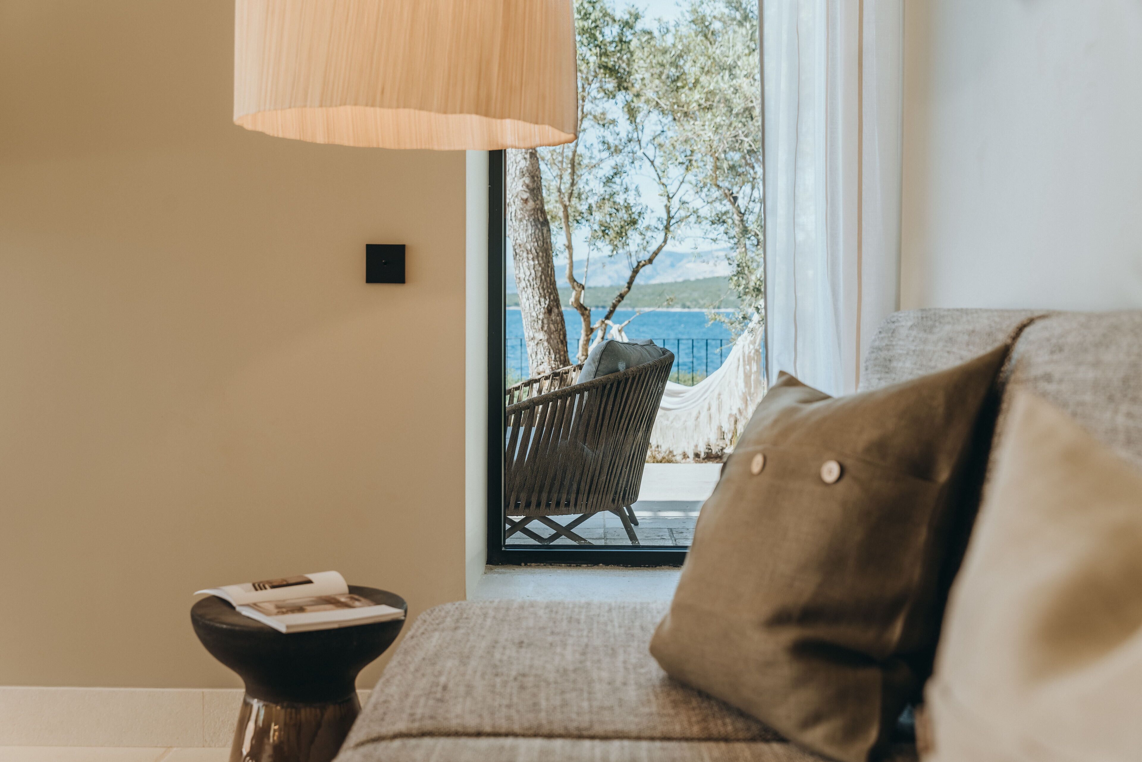 A cozy living room with a view of the sea, a wicker chair, and a side table with a book.