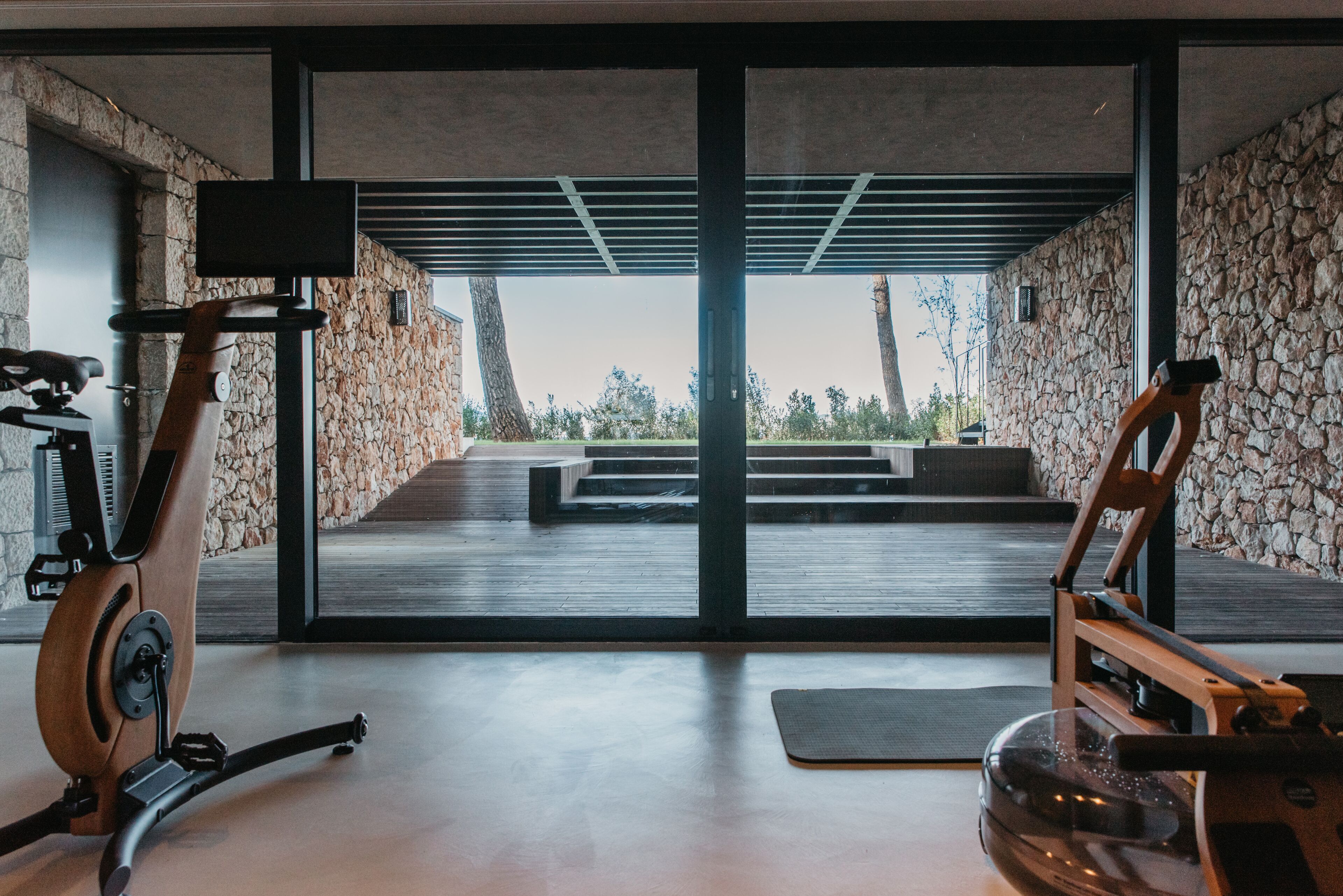 A modern gym with stationary bikes and a large glass door leading to an outdoor area with steps and a view of trees.