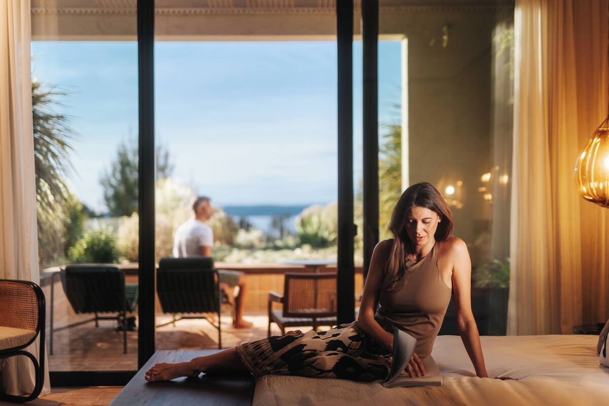 A woman sitting on a bed in a room with a large window overlooking a patio area.