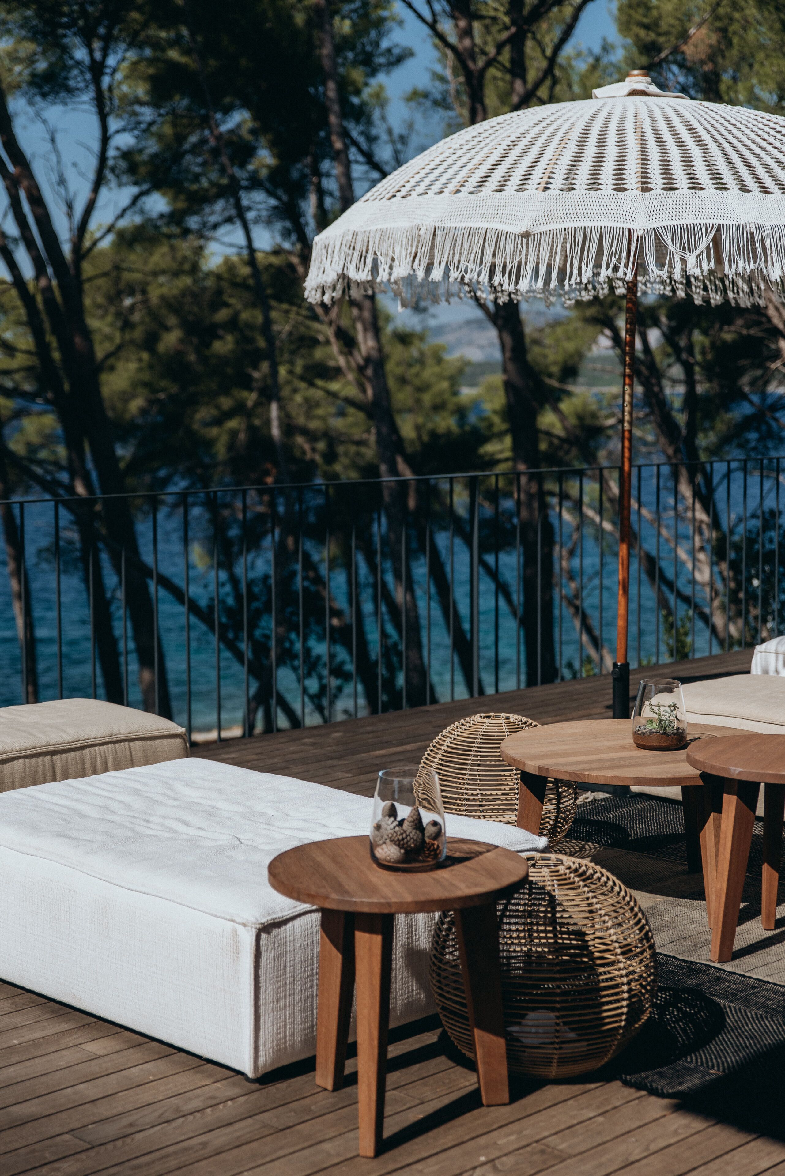 A deck with a white cushioned sofa, round wooden tables, wicker chairs, and a white crocheted umbrella.