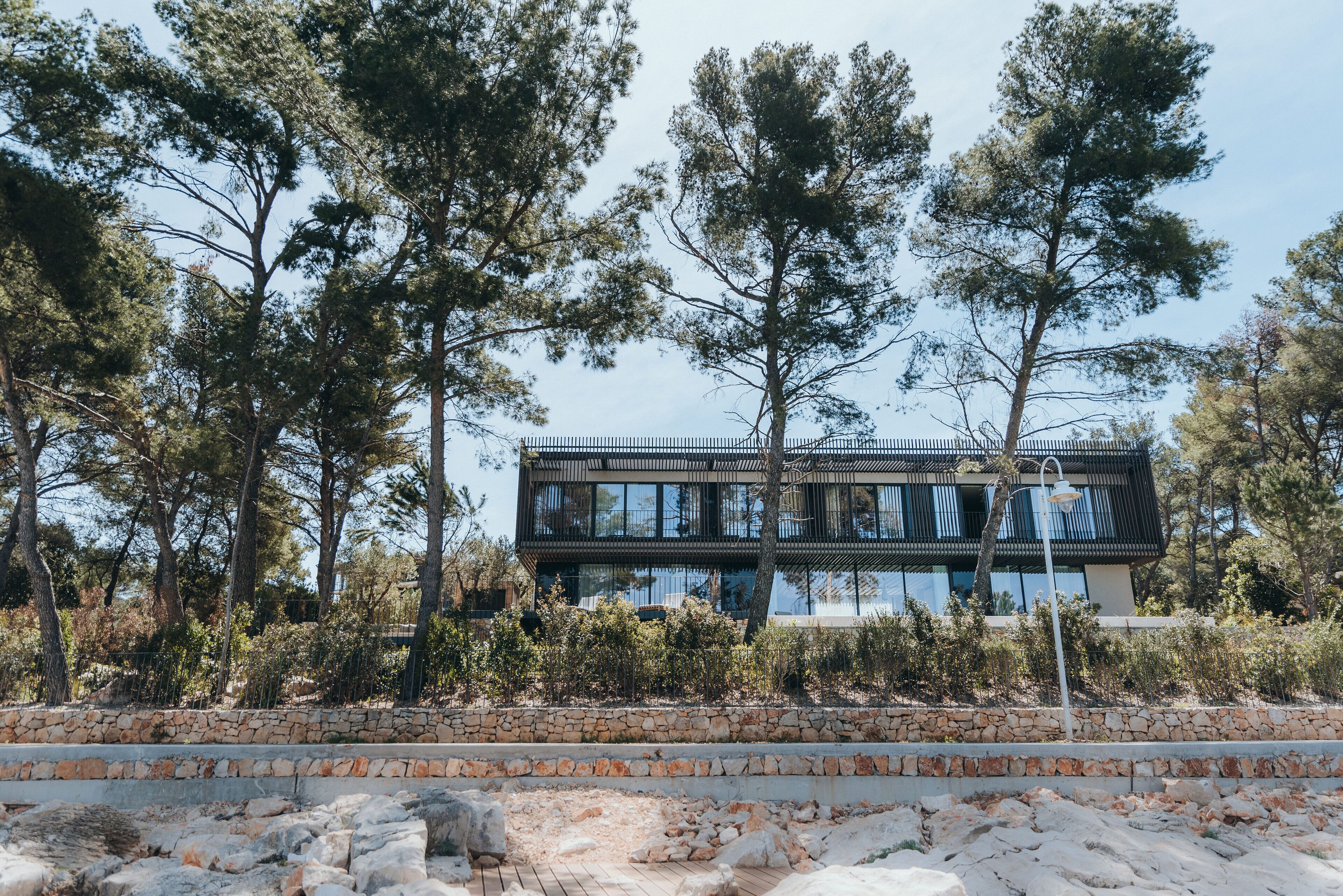 Un edificio moderno de dos pisos con un muro de piedra de contención en el frente, rodeado de árboles y vegetación.