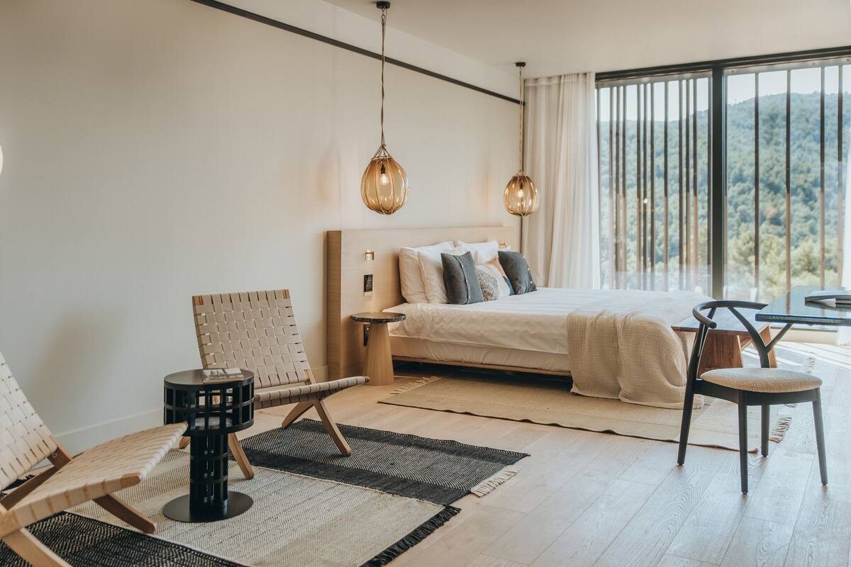 A modern bedroom with a large bed, two side tables, a chair, and a view of the outdoors.