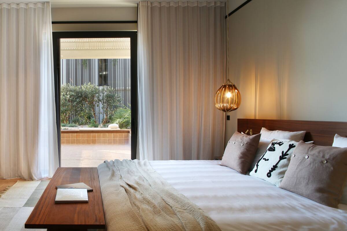 A hotel room with a bed, a wooden side table, and a view of a garden through the curtains.