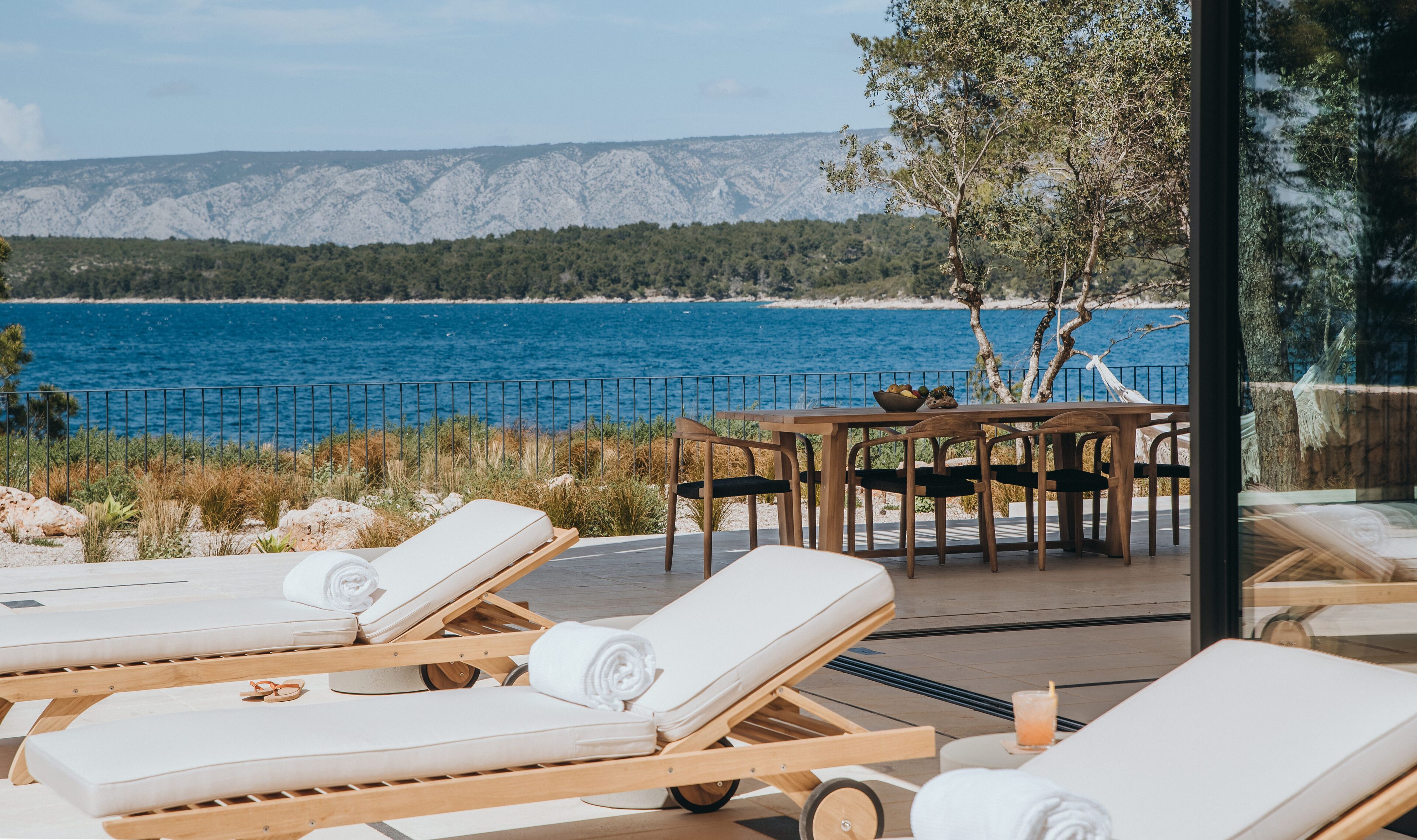 Una terraza frente al mar con tumbonas, una mesa con frutero y vistas al océano y montañas.