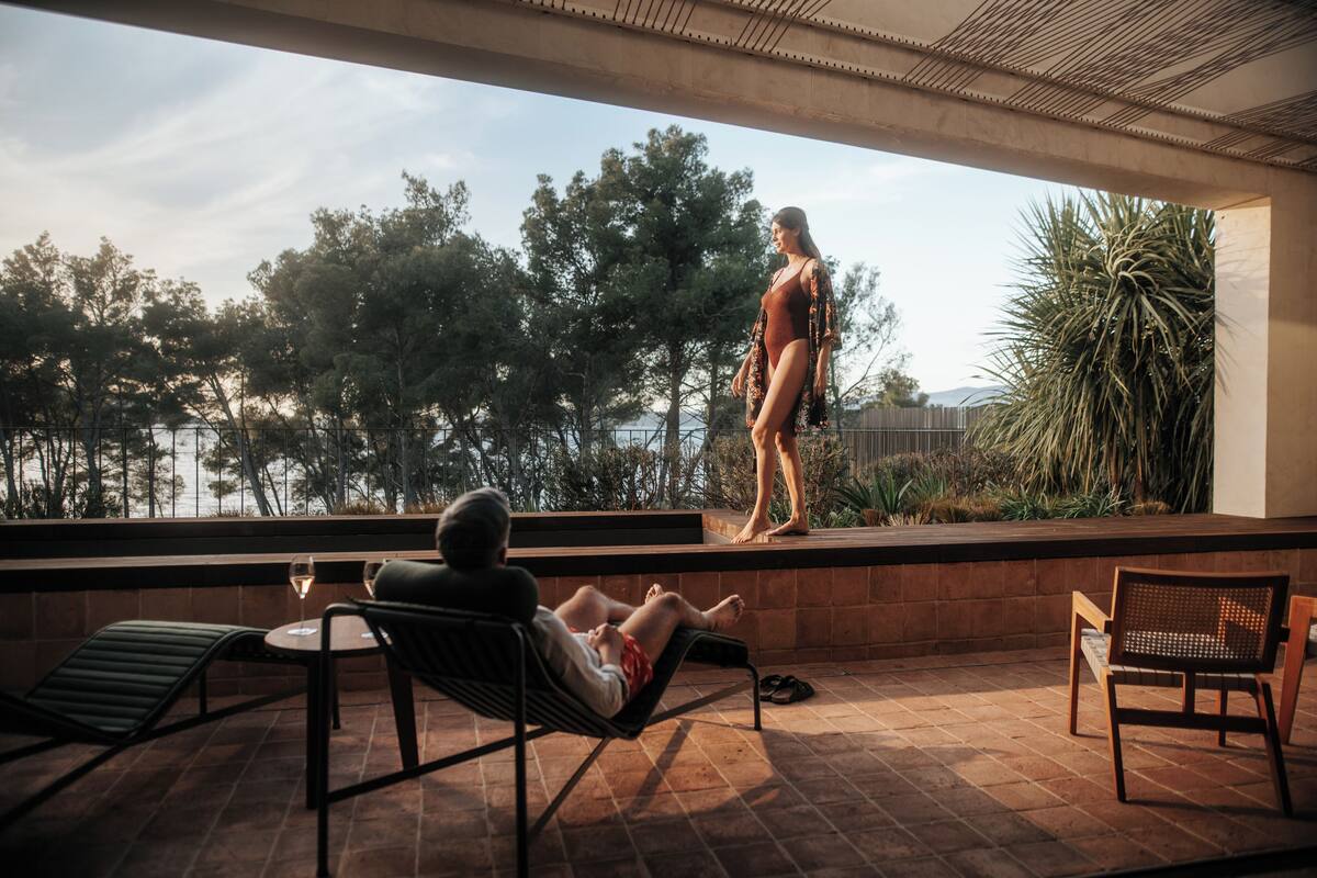 A terrace with a person standing on a platform, a seated individual with a drink, and a chair.
