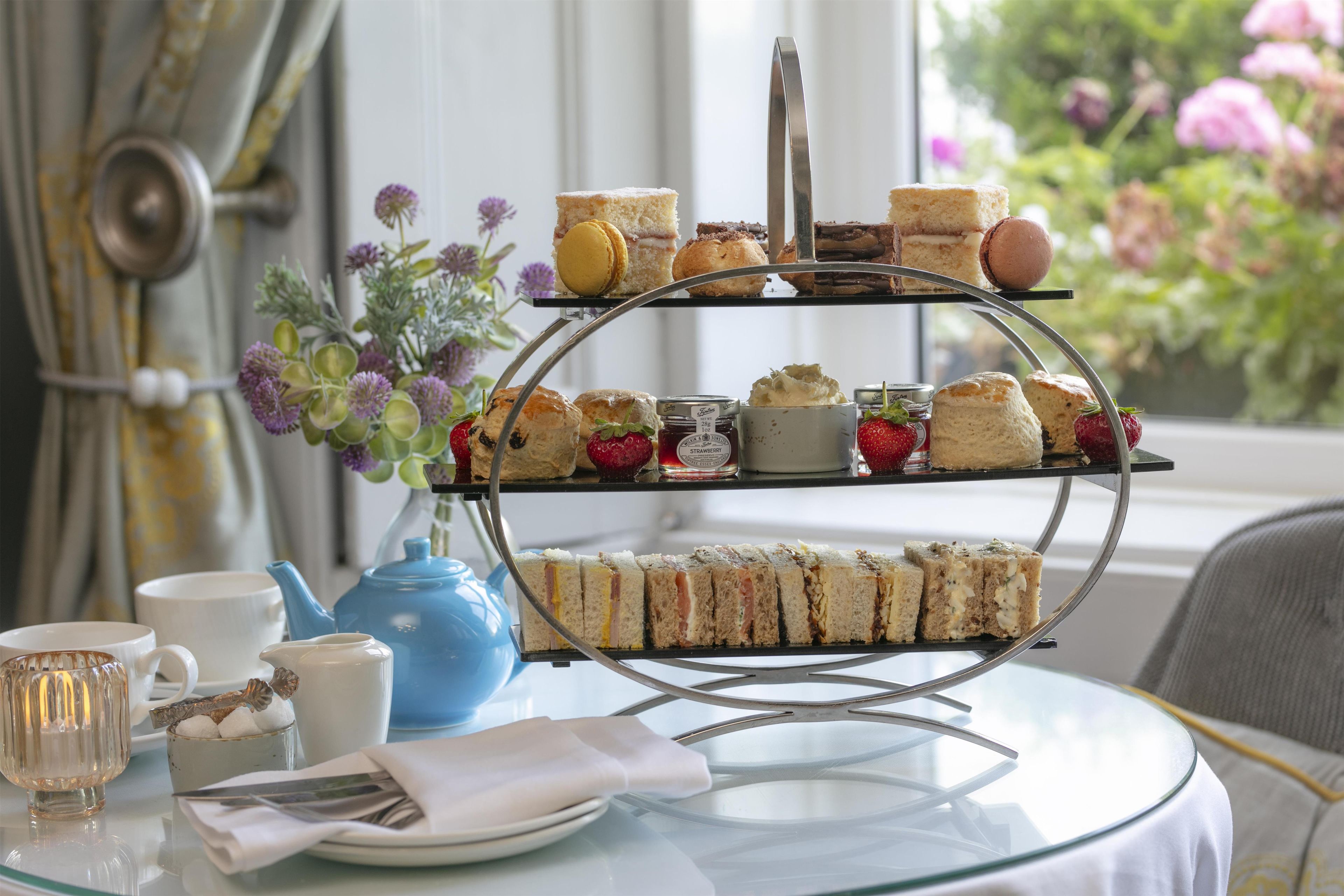 Suporte de sobremesas em três níveis com diversos pastéis e sanduíches, um bule azul e um vaso com flores sobre uma mesa.