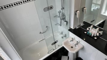 Un baño con ducha envuelta en vidrio, un lavabo blanco y una encimera negra con artículos de aseo.