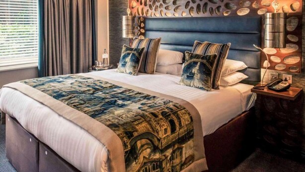 Ein Hotelzimmer mit einem großen Bett, einem dekorativen Kopfteil, einem Nachttisch mit Lampe und Blick ins Freie durch das Fenster.
