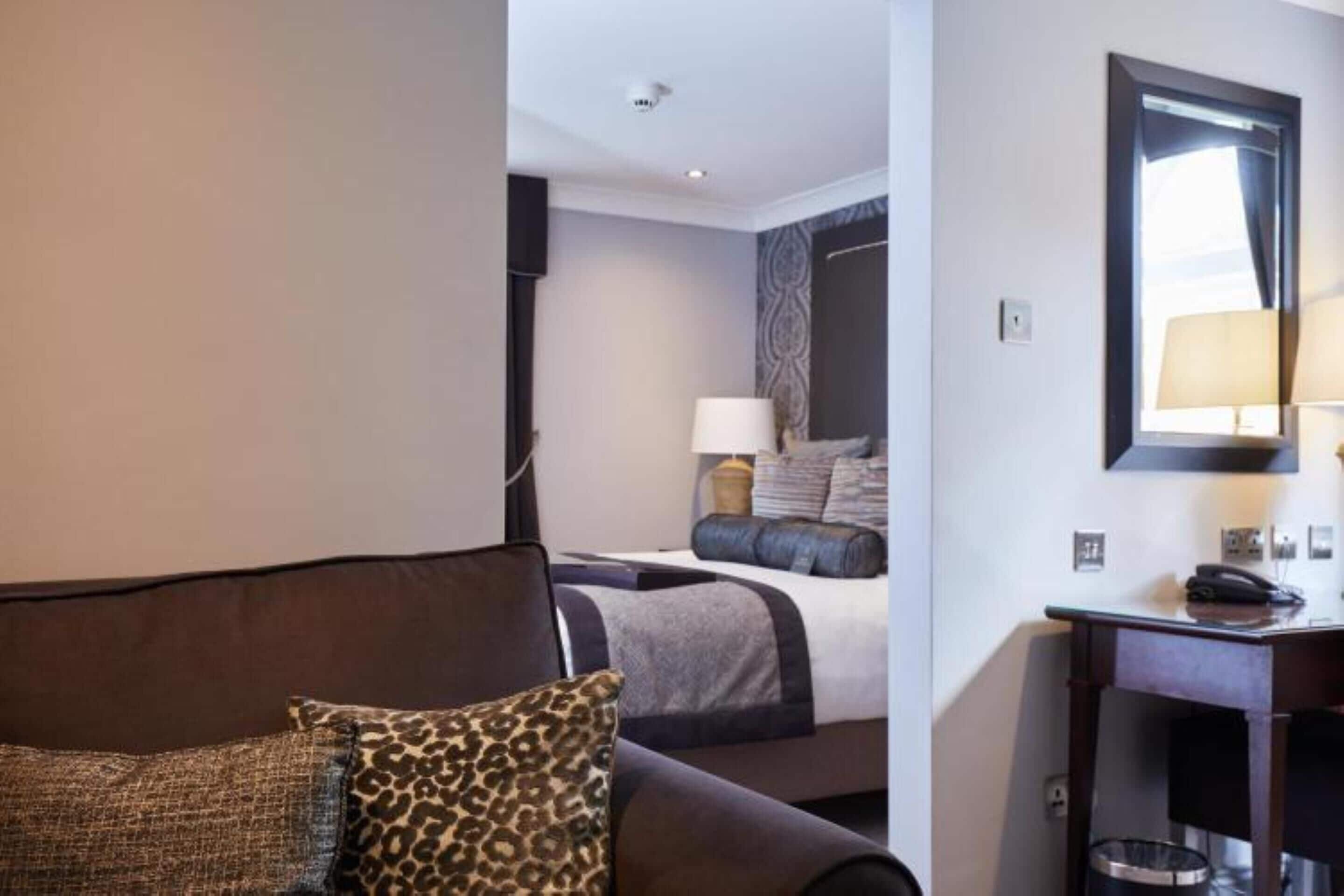 Une chambre d’hôtel avec un canapé marron, un lit doté d’une tête de lit à motifs, une table de chevet avec une lampe et un miroir au mur.