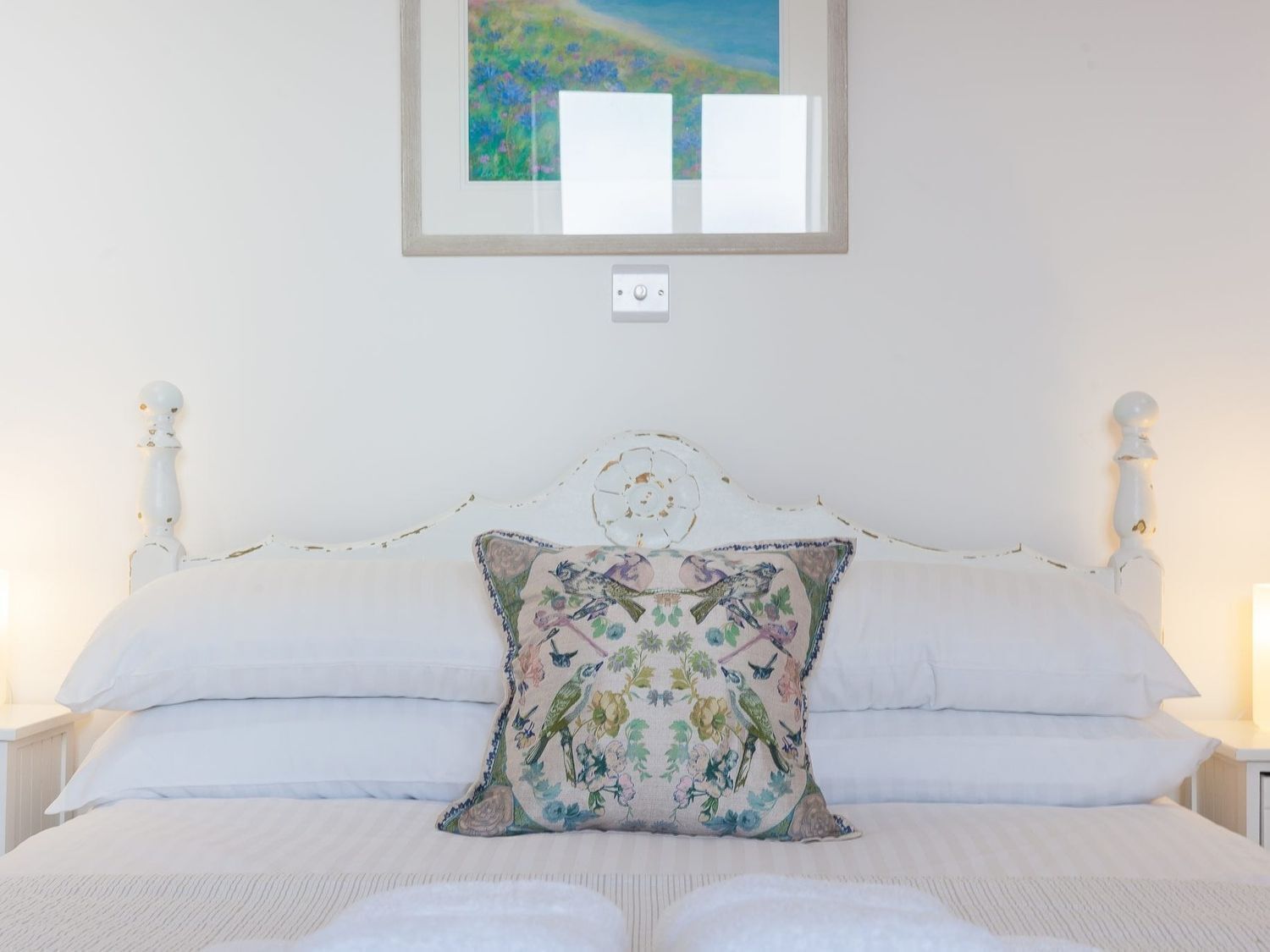 Une chambre avec un lit blanc, un coussin à motifs floraux et un tableau accroché au-dessus du lit.