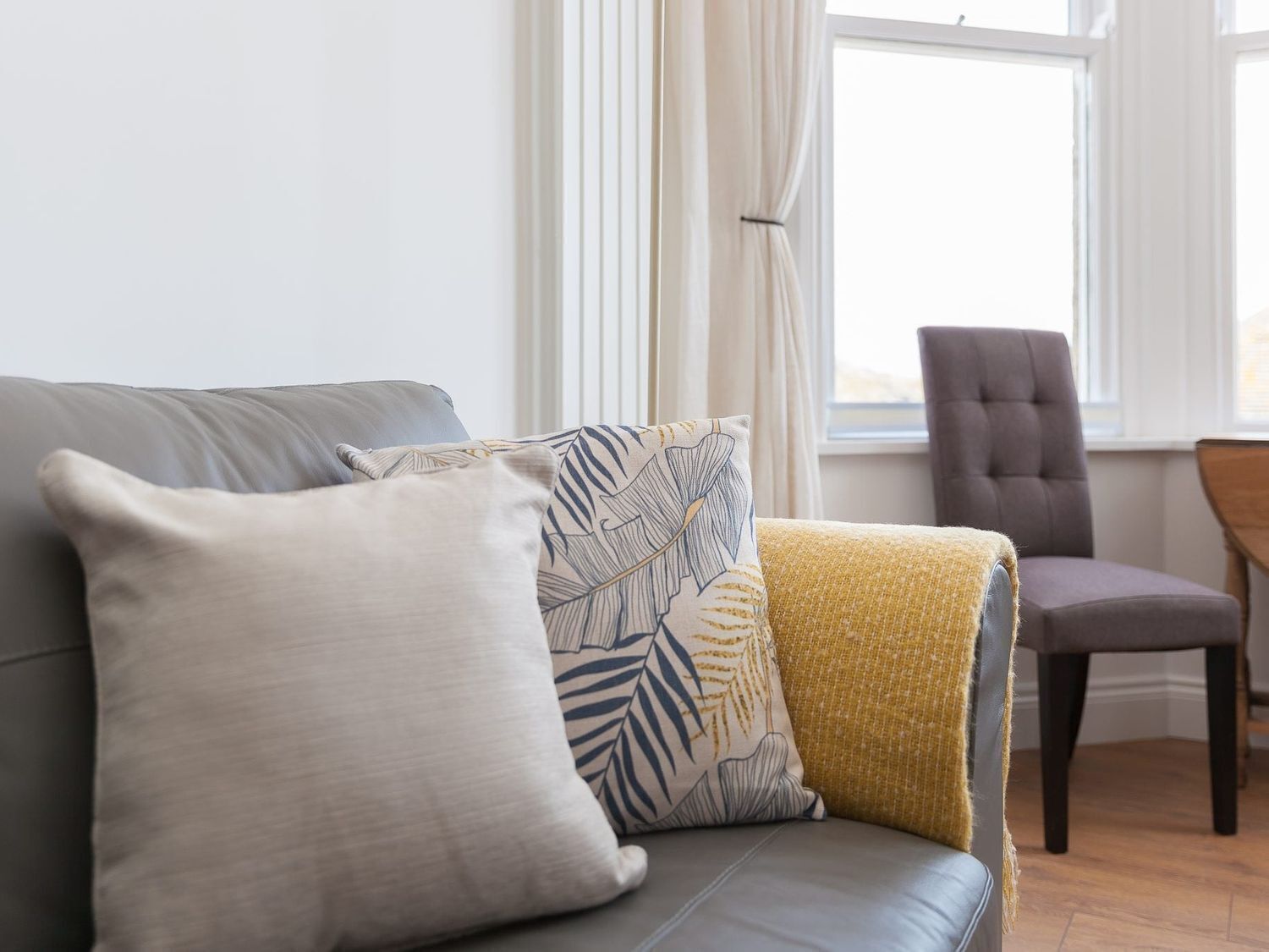 A grey sofa with three cushions, a yellow throw blanket, and a dark brown chair in a room with a window and curtains.