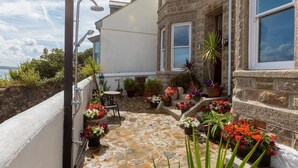 Ein Steinhaus mit weißer Fassade, eine kleine Terrasse mit Tisch und Stühlen sowie verschiedene Kübelpflanzen.