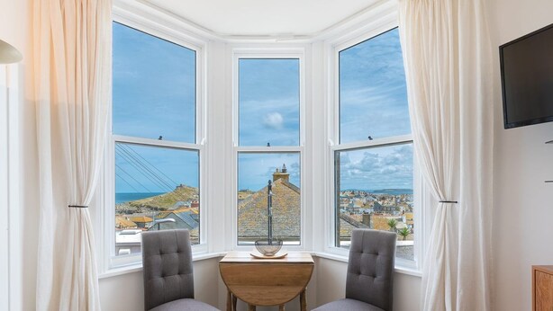 Ein Eckzimmer mit drei Erkerfenstern mit Blick auf eine Küstenstadt, ausgestattet mit einem Holztisch und zwei grau gepolsterten Sesseln.
