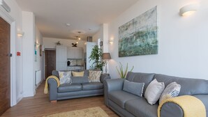 Ein modernes Wohnzimmer mit einem grauen Sofa, einem kleineren grauen Sofa und einem gelben Teppich. Im Hintergrund ist eine Küche mit weißen Schränken und einer grünen Pflanze zu sehen.