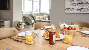 Een ontbijttafel gedekt met croissants, fruit en sapglazen, met uitzicht op een zithoek en een muurschildering van vissen op de muur.