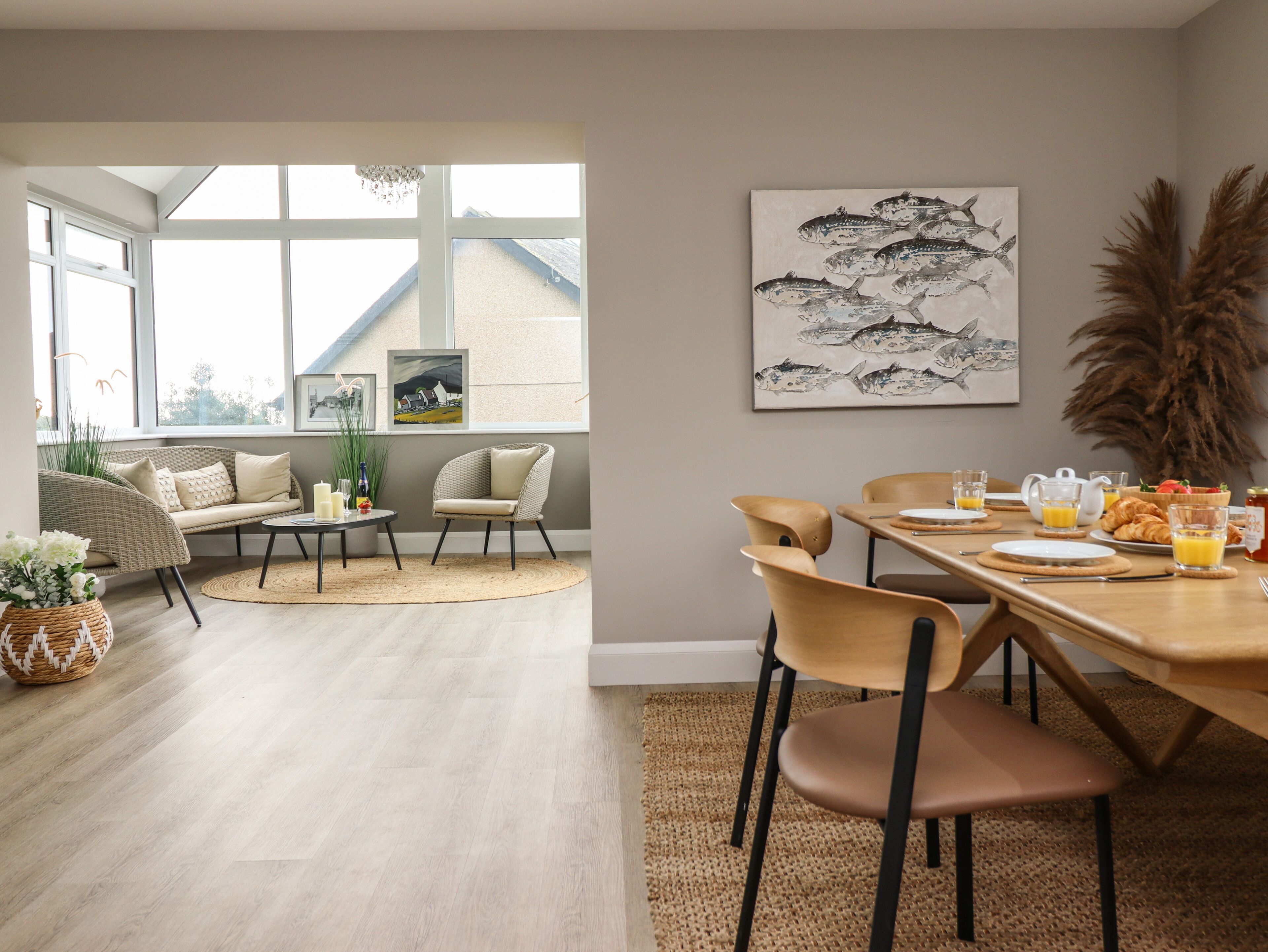 Un comedor moderno con un gran ventanal, mobiliario de mimbre y una mesa de desayuno servida.