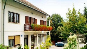 Un edificio de dos pisos con un balcón decorado con flores rojas, un letrero que dice "Zimmer Rooms" y un pequeño espacio al aire libre con mesas y sillas.