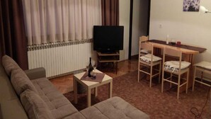 Una sala de estar con sofá, una mesa de centro con una botella y vasos, televisor, mesa de comedor con sillas y un radiador.