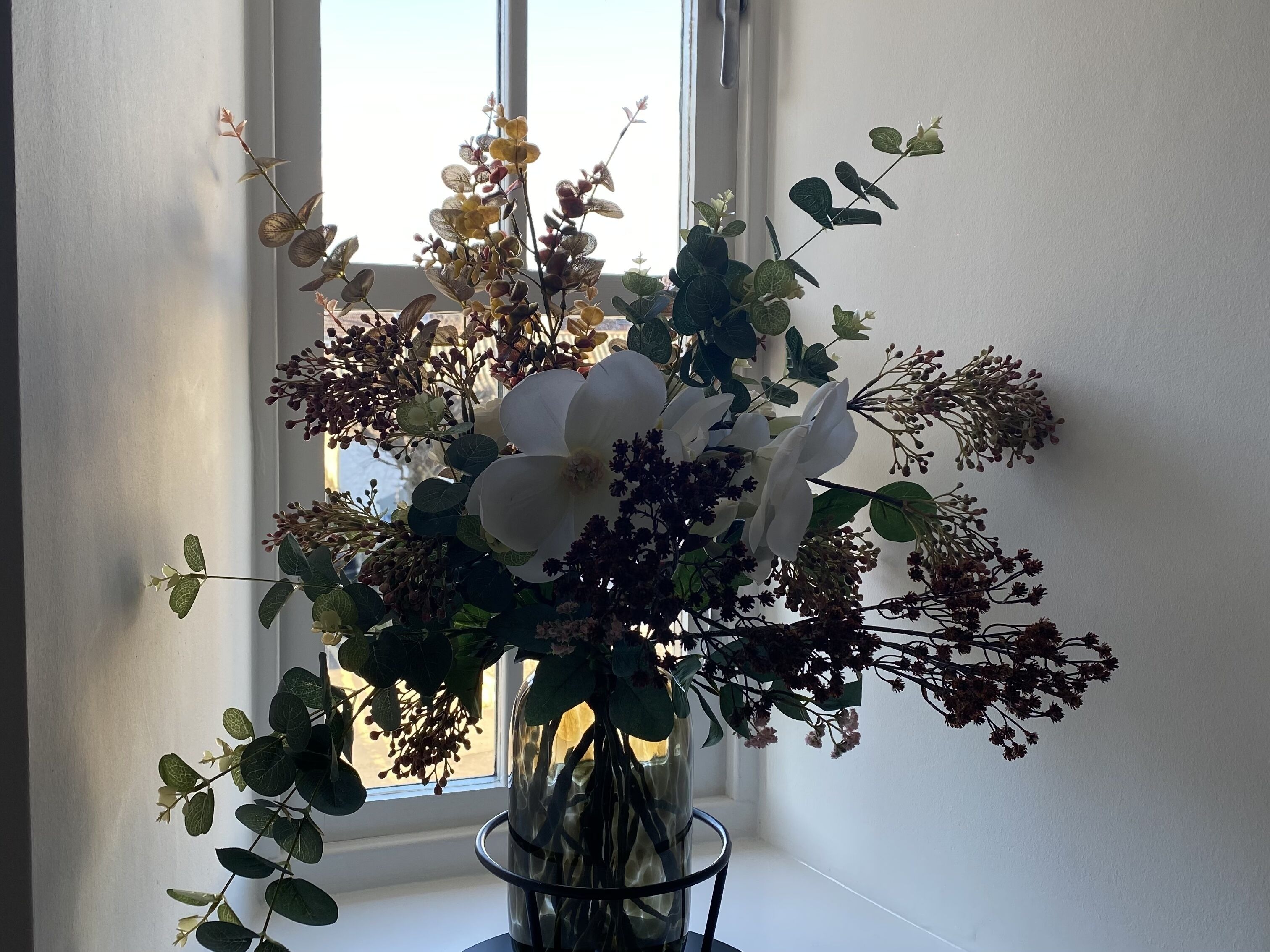 Een vaas met verschillende bloemen en groen op een vensterbank.