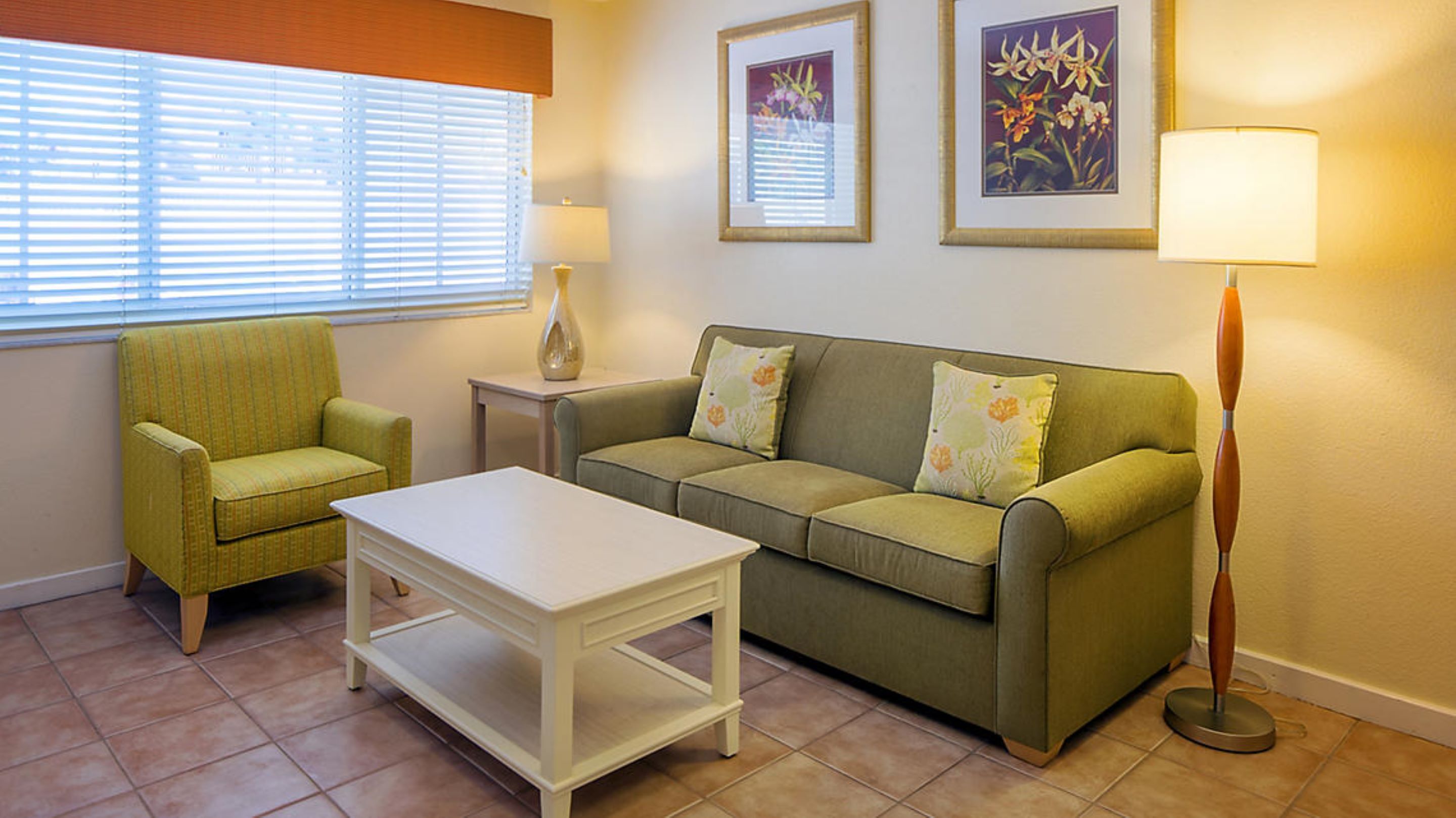 Un salon avec un canapé vert, un fauteuil assorti, une table basse blanche et un lampadaire. Deux tableaux encadrés sont accrochés au mur.
